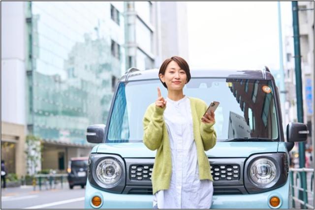 左手にスマホを持ち、右手の人さし指を立てて車の前に立つ女性
